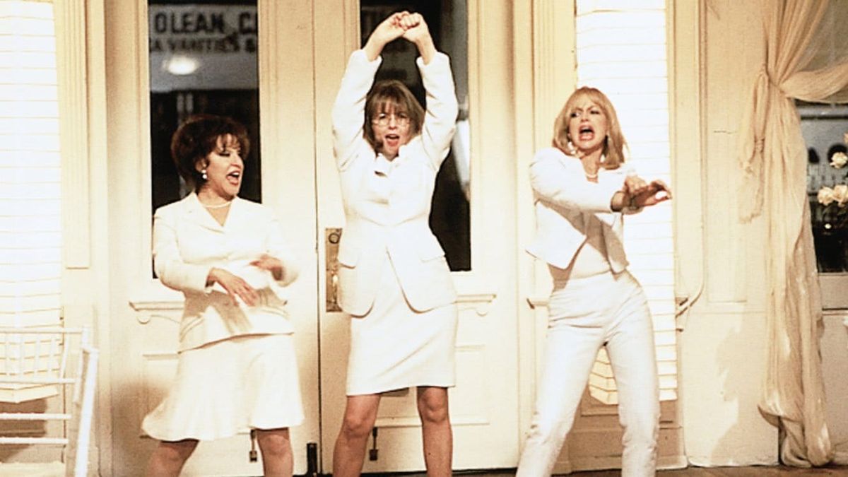 Three women in white outfits dancing energetically on a porch, striking fun poses with arms raised and extended.