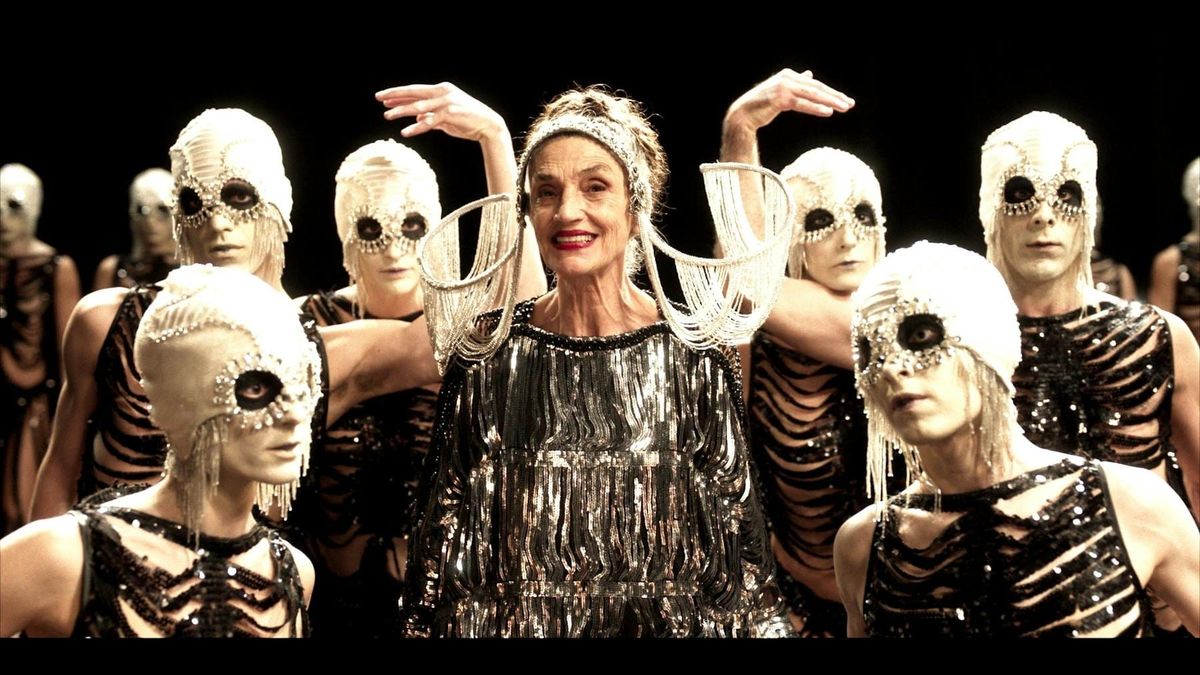 Elderly woman in sparkly fringed dress with arms raised, surrounded by performers in skeletal costumes and skull-like masks.