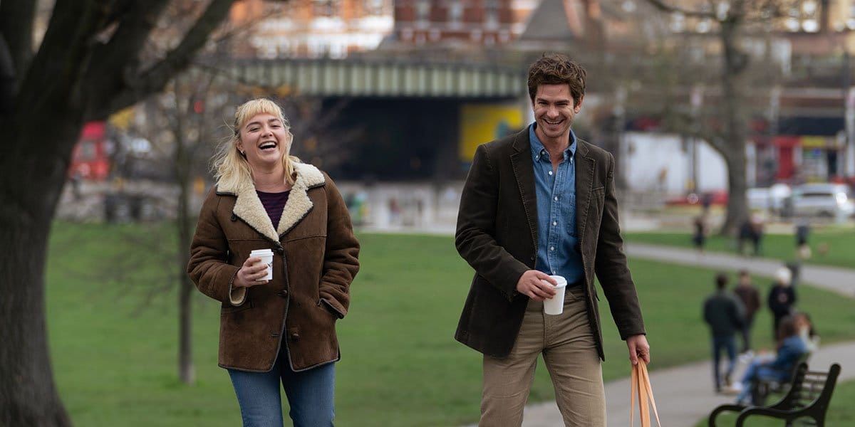 Two people laughing while walking through a park holding coffee cups, both wearing brown jackets on an overcast day.