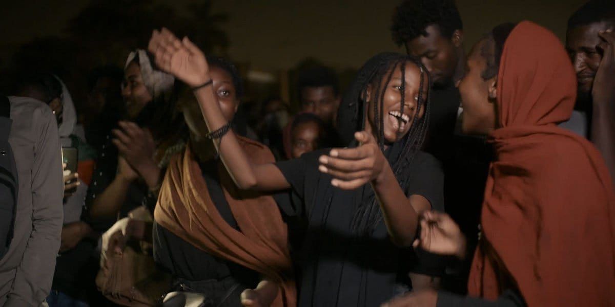 Young people dancing and laughing together at a nighttime gathering, with a woman in braids reaching out joyfully.