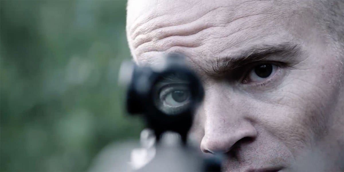 Close-up of a bald man aiming a rifle with a scope, one eye looking through the sight, outdoors with blurred green background.
