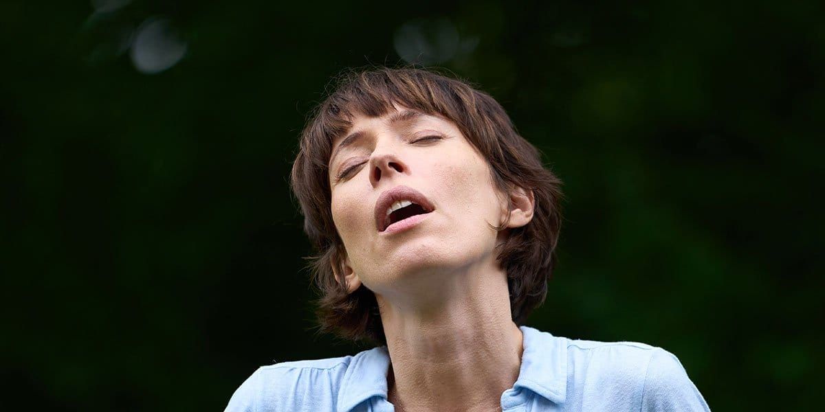 Woman with short brown hair and closed eyes tilting her head back with mouth open, wearing a light blue shirt, outdoors.