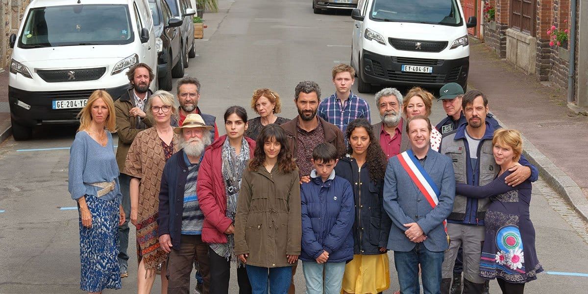 Group of about 18 people standing together on a French village street, one wearing a tricolor mayoral sash, cars parked behind.
