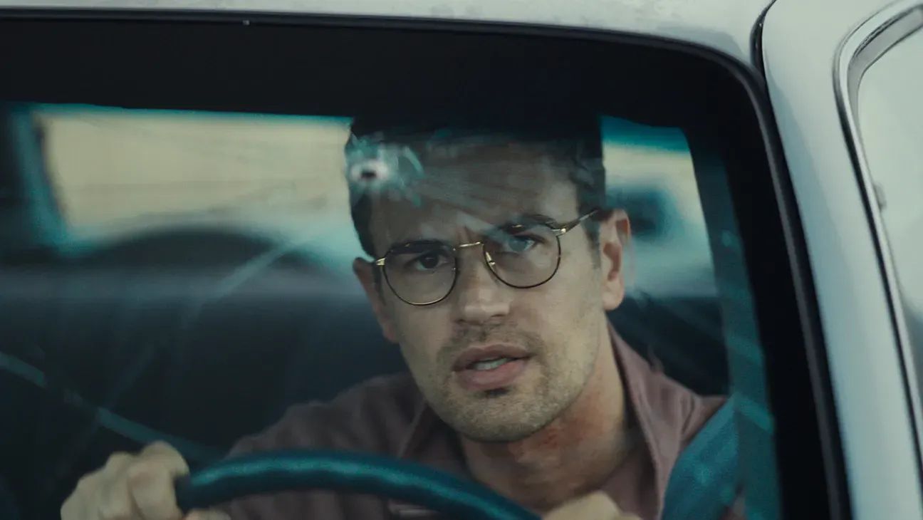Man wearing round glasses sits behind steering wheel of a car, looking concerned, with a bullet hole in the windshield.