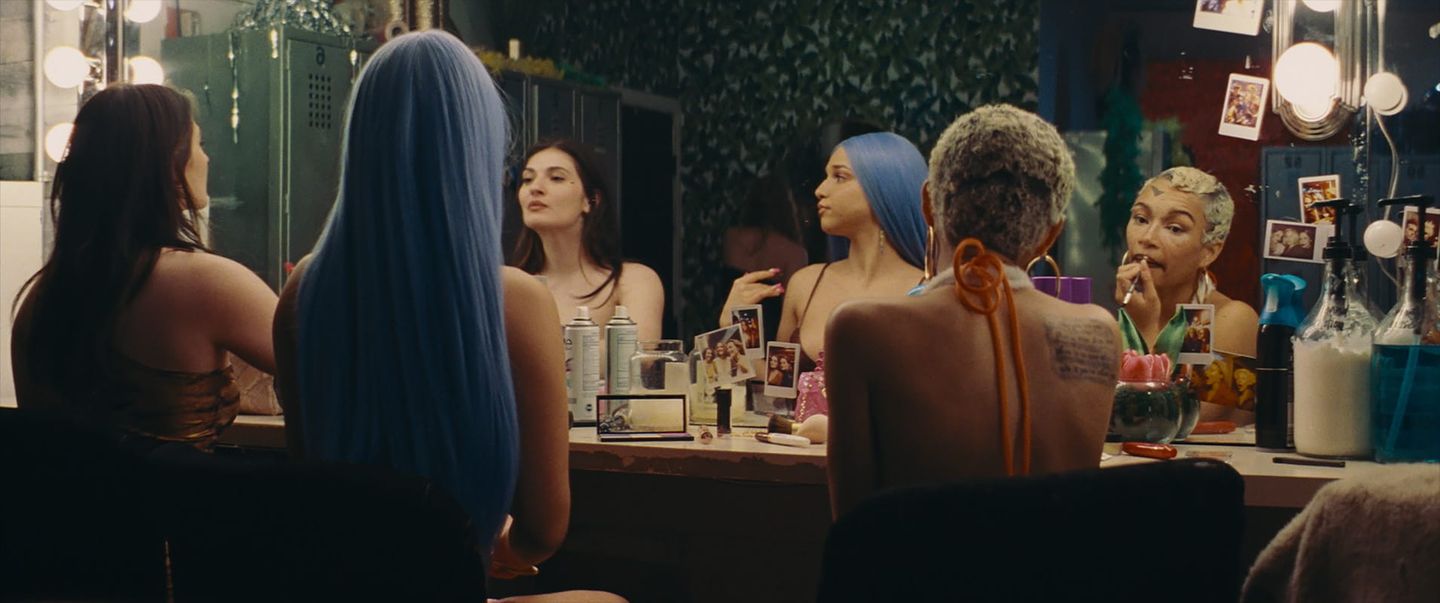 Three women sit at a backstage dressing room vanity with lit mirrors, applying makeup, wearing colorful wigs and costumes.
