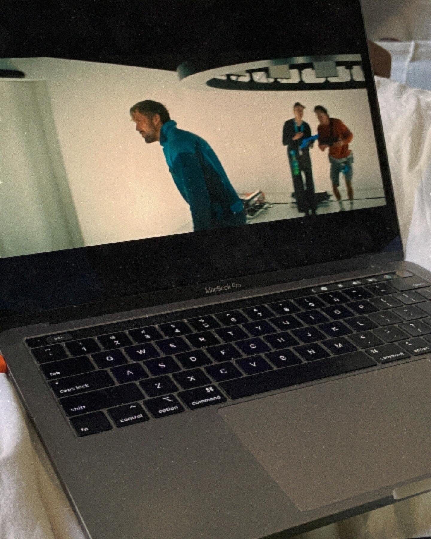 MacBook Pro displaying a movie scene with a man in a blue jacket on a futuristic set, two people visible in background.