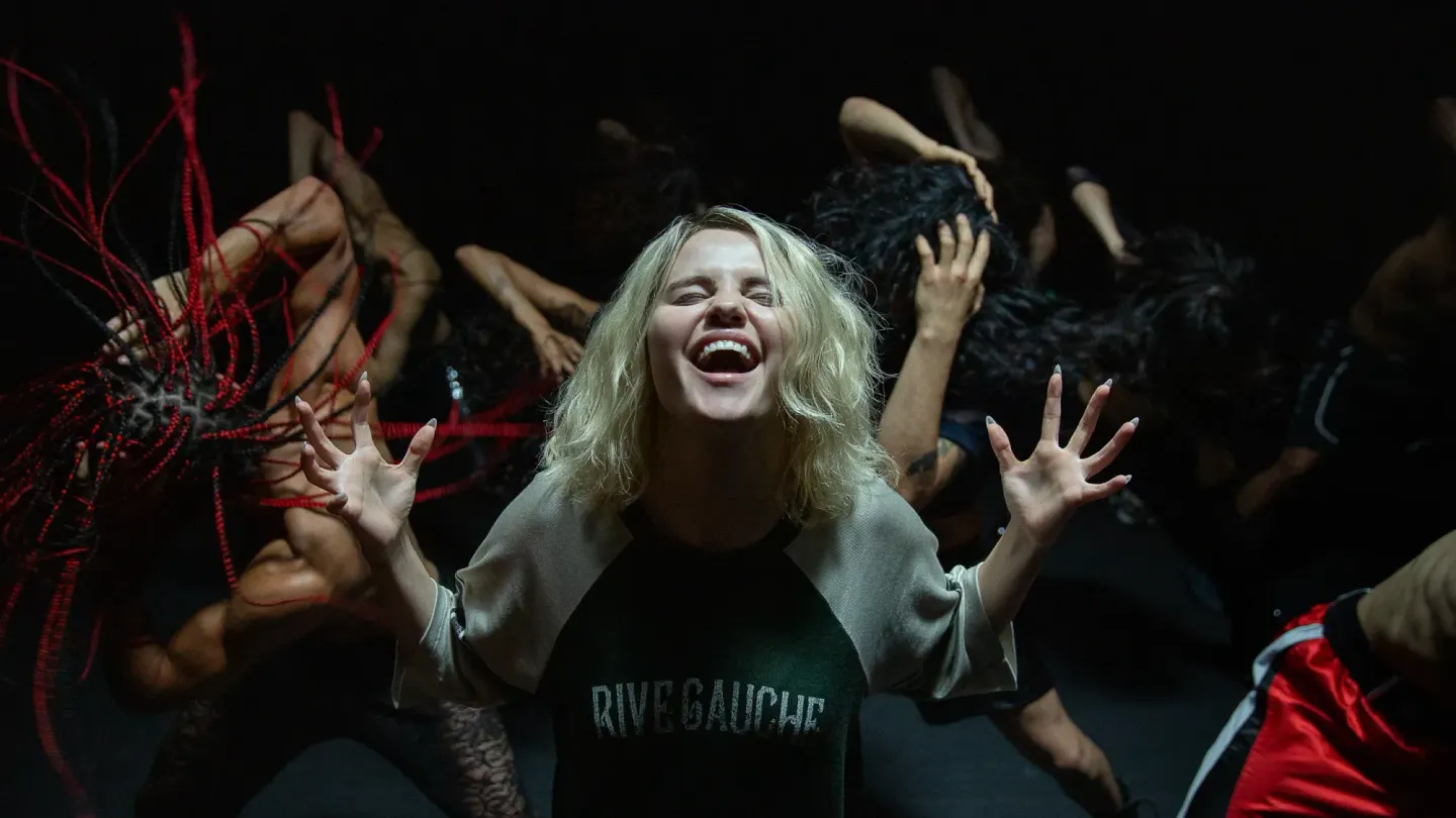 Blonde woman in "Rive Gauche" shirt laughing with outstretched hands, surrounded by dancers in a dark performance space.