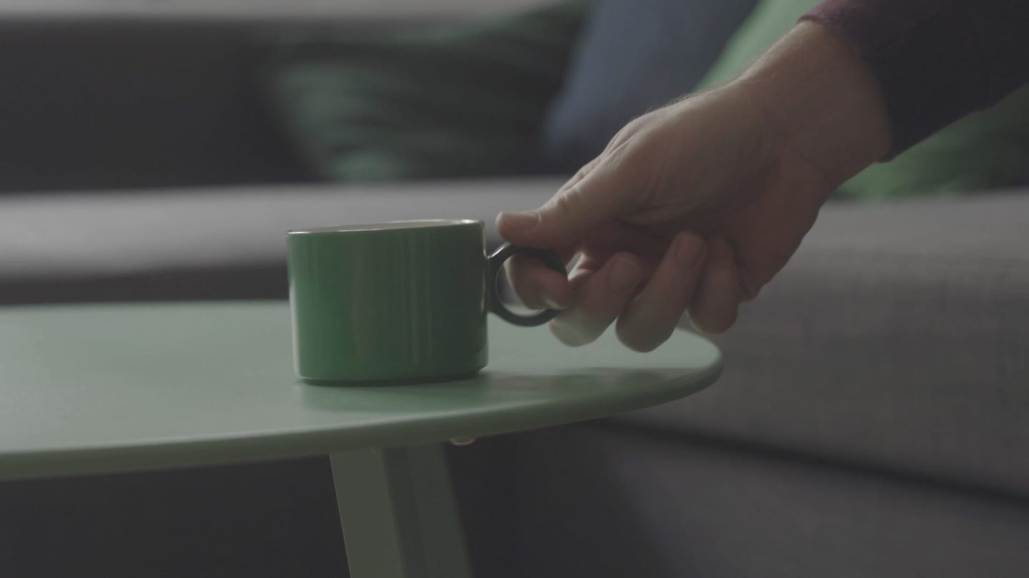 Hand reaching for a green ceramic mug on a sage green side table, with a gray couch in the blurred background.