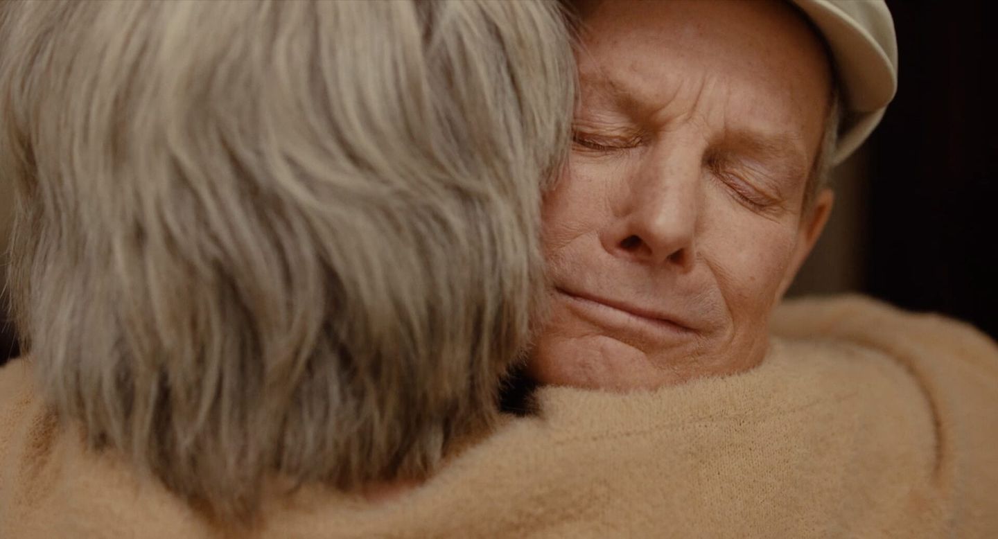 Two elderly people sharing an emotional embrace, one with eyes closed and a gentle smile, the other with gray hair facing away.