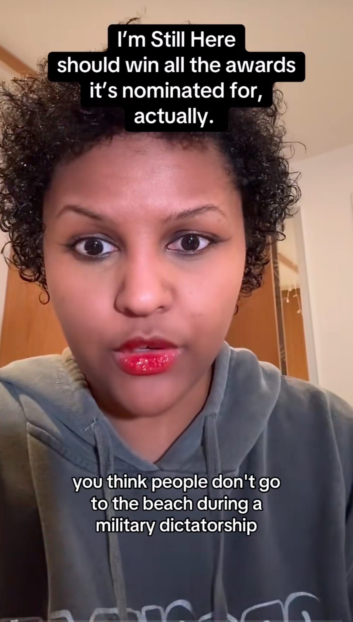 Woman with curly hair and red lipstick in gray hoodie, with text overlay praising film "I'm Still Here" and discussing beach during military dictatorship.