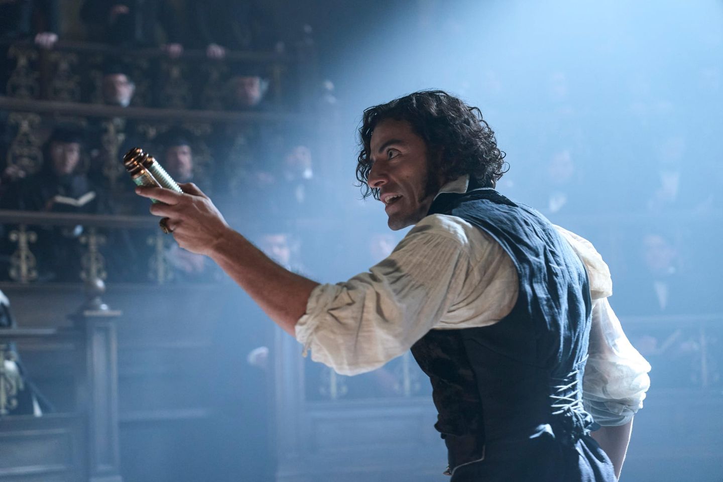 Actor in period costume on stage holding a small object aloft, with audience visible in balcony seating behind hazy lighting.