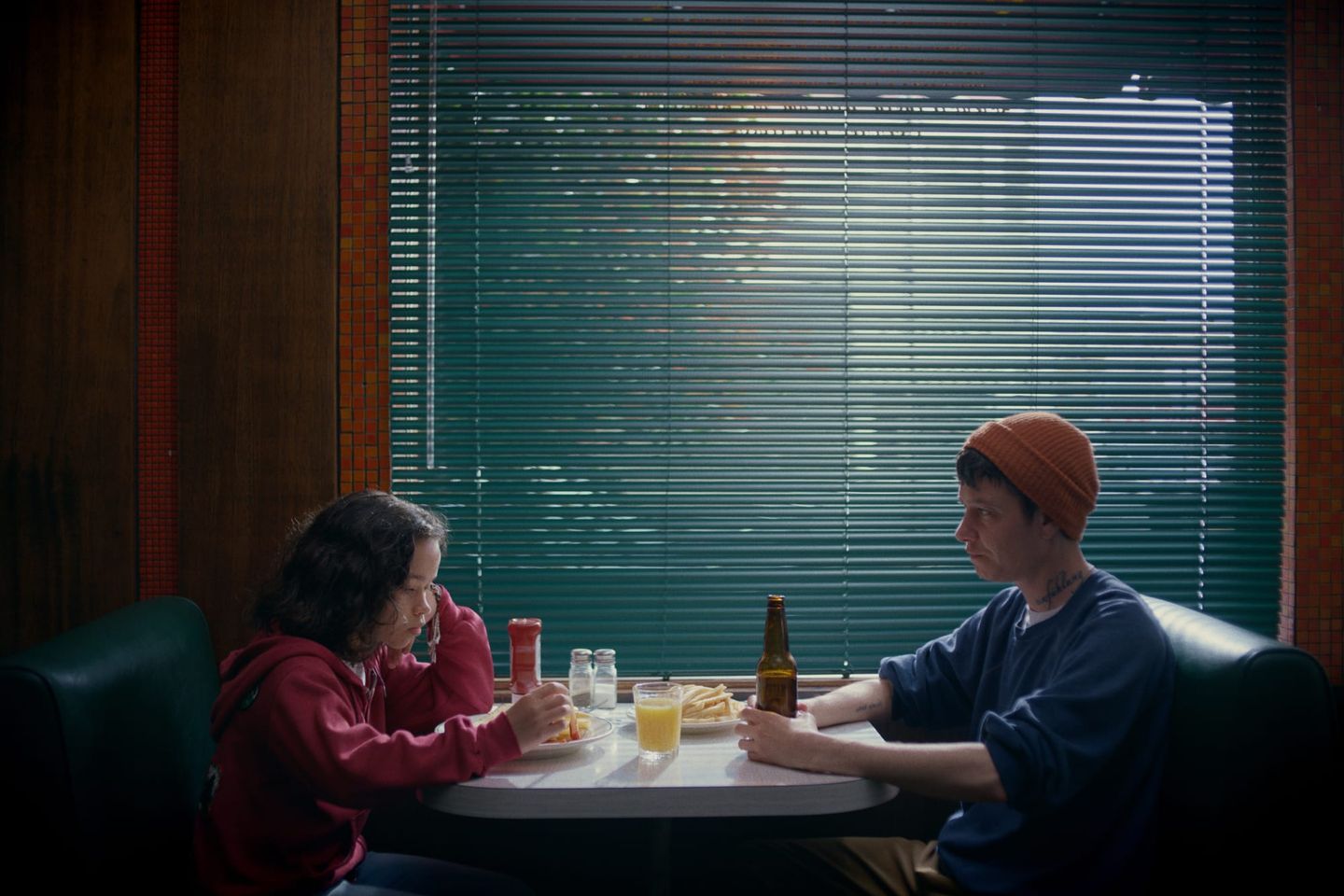 Two people sit across from each other in a diner booth eating fries and drinking, with venetian blinds behind them.