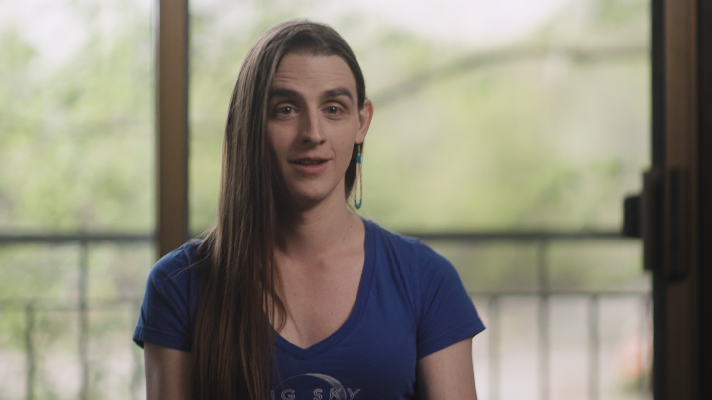 Person with long brown hair wearing a blue "Big Sky" t-shirt and turquoise earring, speaking indoors near large windows.