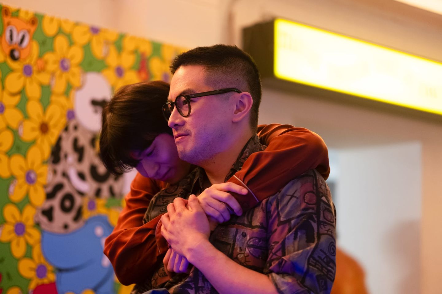 Child hugs a man wearing glasses and a patterned shirt from behind, clasping hands, in front of a colorful cartoon mural.