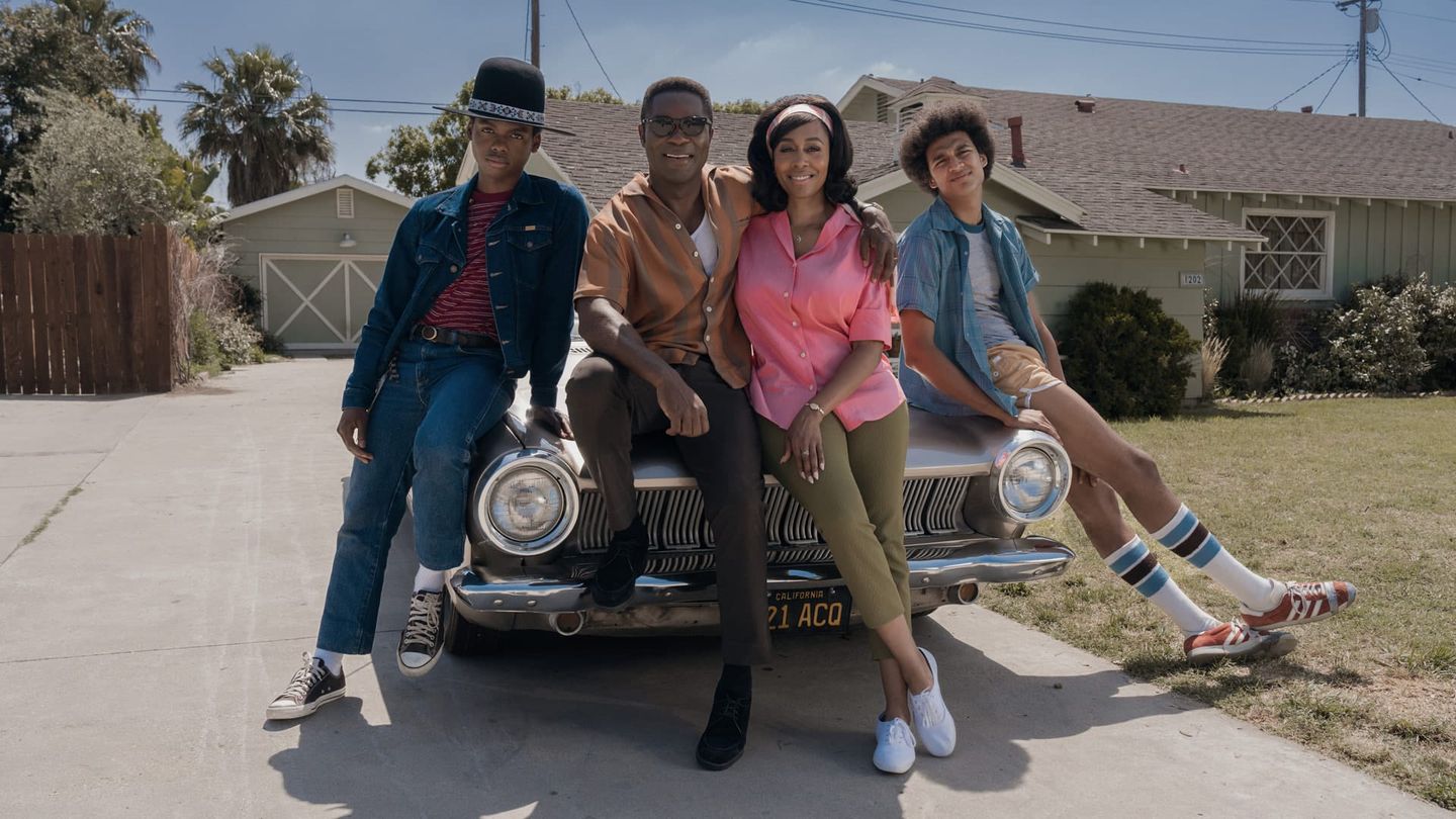 Four people sitting on the hood of a vintage car with California plates in a suburban driveway, dressed in 1970s attire.
