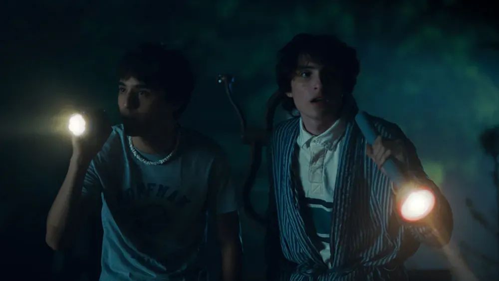 Two teen boys holding flashlights in a dark outdoor setting, looking alarmed, in a cinematic horror movie scene.