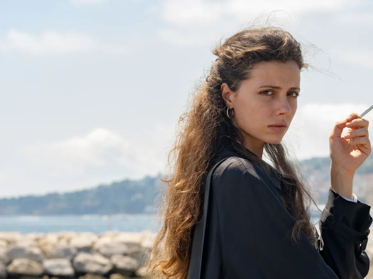 Young woman with long wavy brown hair in black blazer holding a cigarette, looking over her shoulder near a rocky coastline.