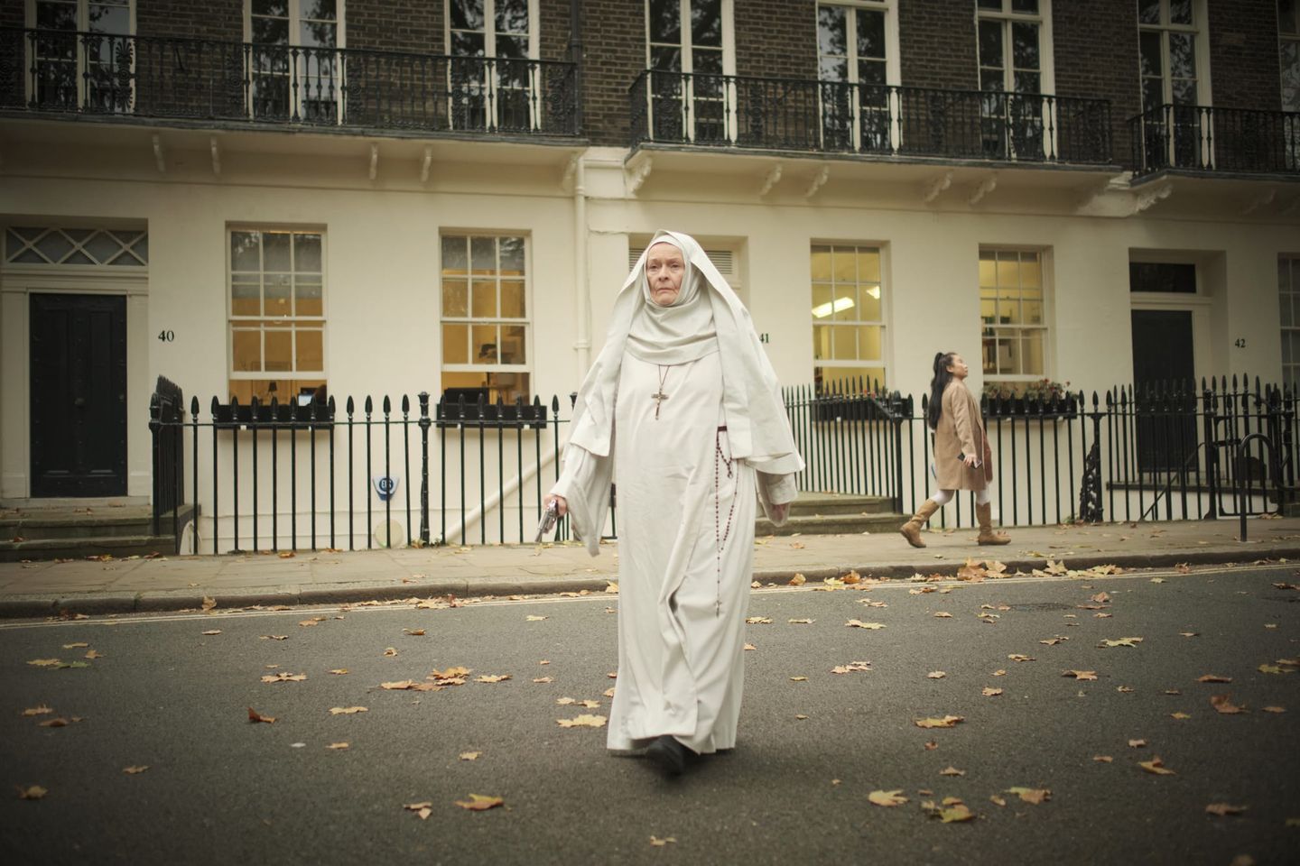 Elderly nun in white habit with crucifix and rosary walks across a leaf-strewn London street near buildings numbered 40-42.