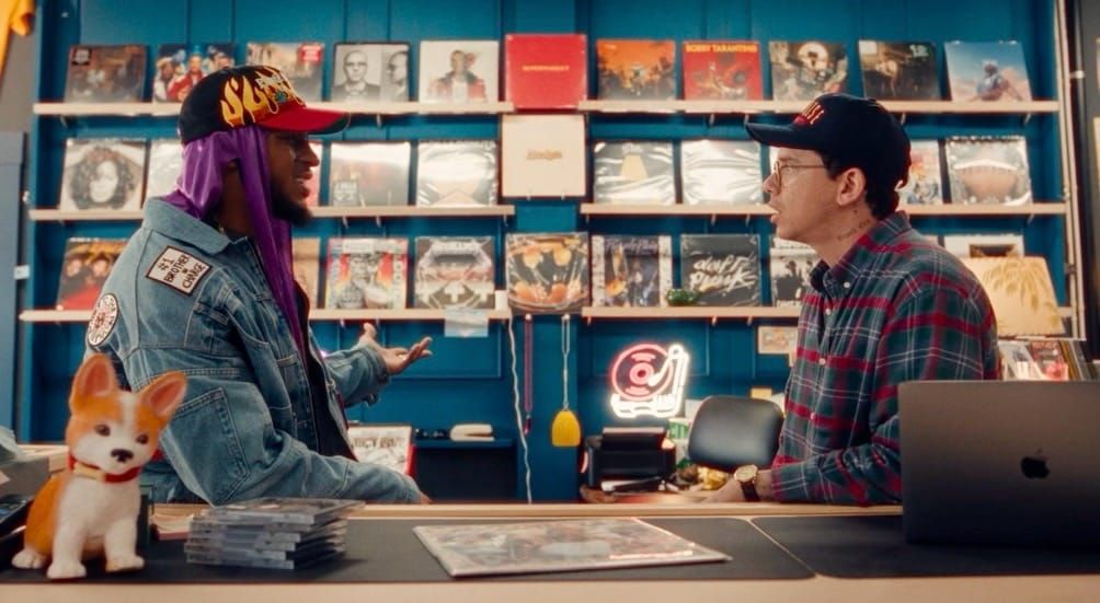 Two men talking at a record store counter, one in a denim jacket with purple durag, the other in plaid shirt and cap.