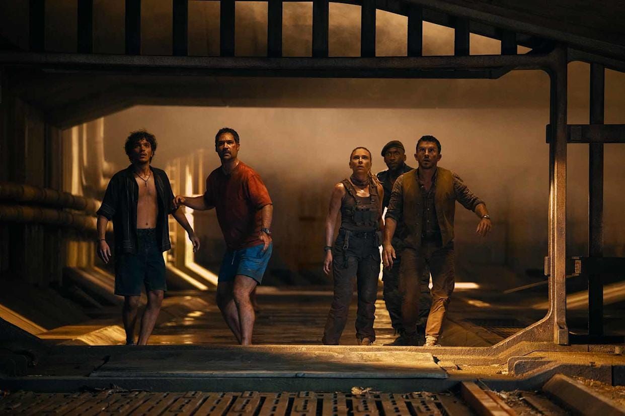 Five people walk cautiously through a dim, industrial corridor with pipes and metal grating, looking tense and alert.