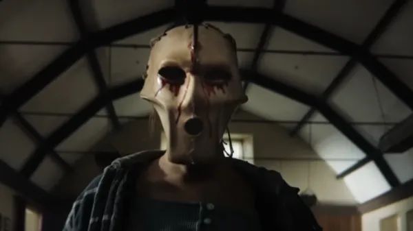 Person wearing a crude, stitched-together leather mask with dark eye holes and blood stains, standing in a dimly lit room.