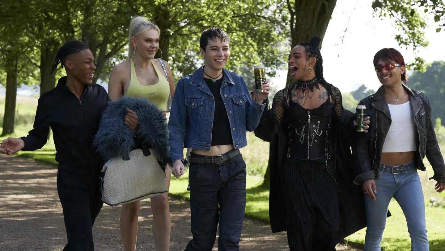 Five young friends walking arm-in-arm on a tree-lined path, laughing and holding canned drinks on a sunny day.