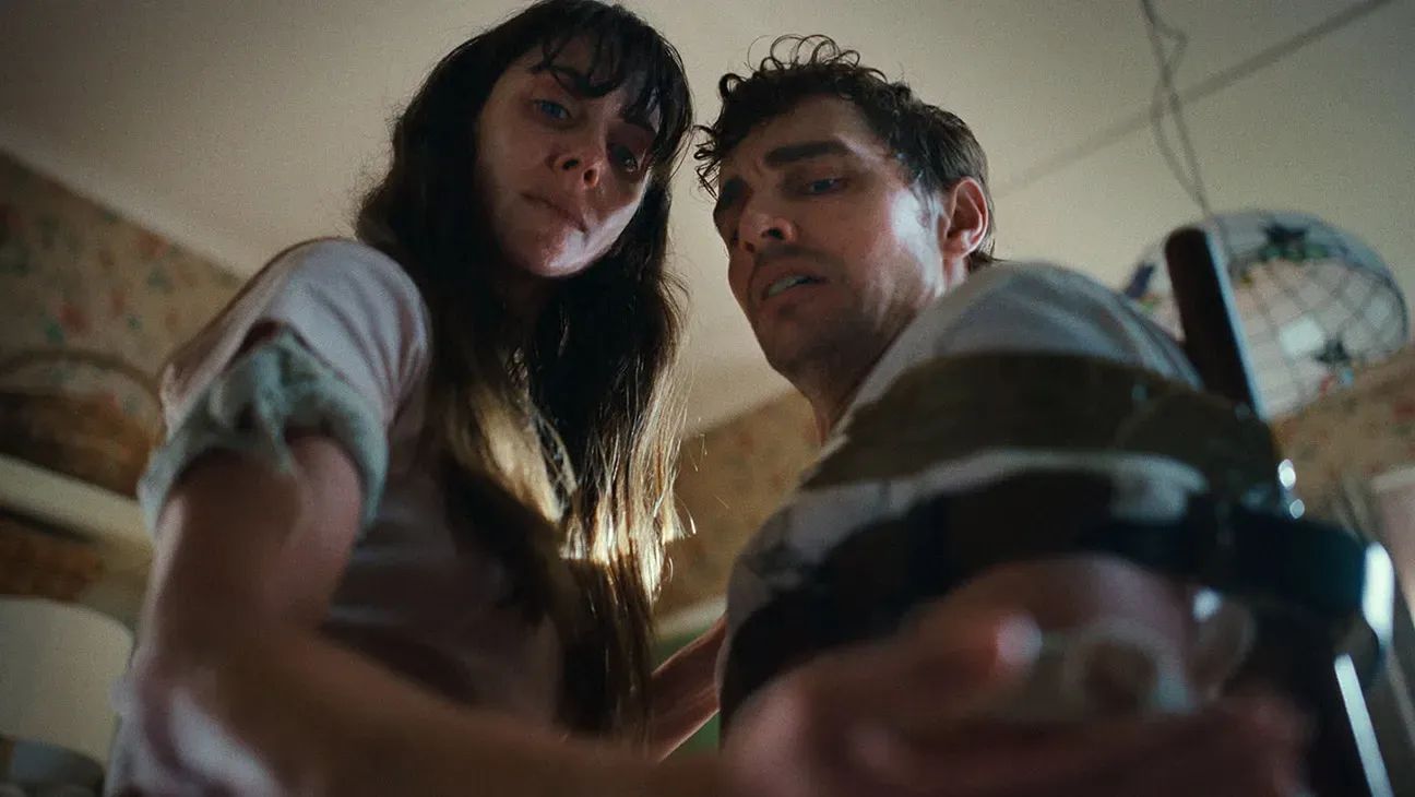 Two people looking down at a glass jar from a low angle in a dimly lit room, appearing curious or concerned.