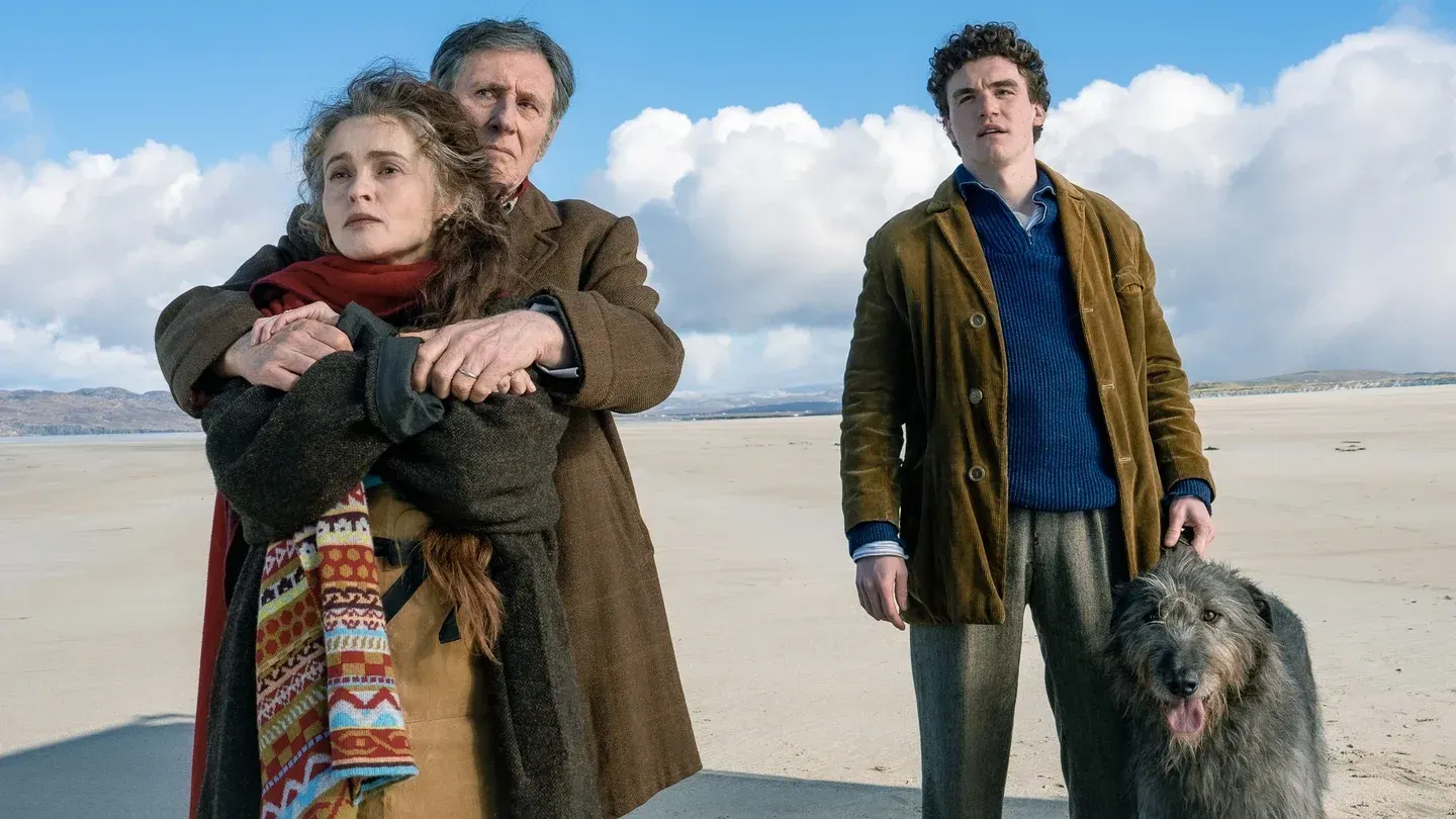 Three people on a windswept beach: a woman in a red scarf embraced by an older man in a brown coat, a young curly-haired man with a large shaggy dog.