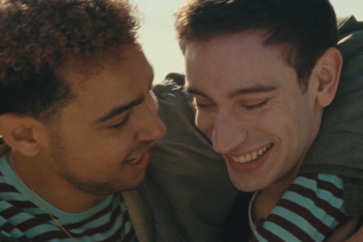 Two young men in an intimate, affectionate moment, faces close together, one smiling with eyes closed, warm golden light.