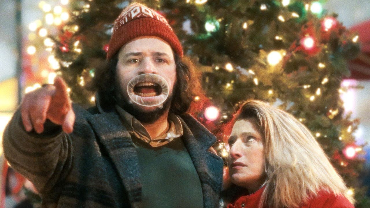 Man in red beanie pointing and yelling with a mouth retractor, woman in red coat beside him, lit Christmas tree behind.