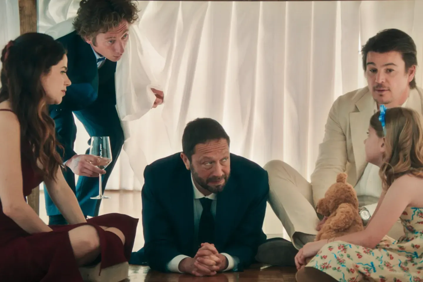 Five people huddle under a table with white draping; a man in a suit sits center, flanked by adults and a girl with a teddy bear.