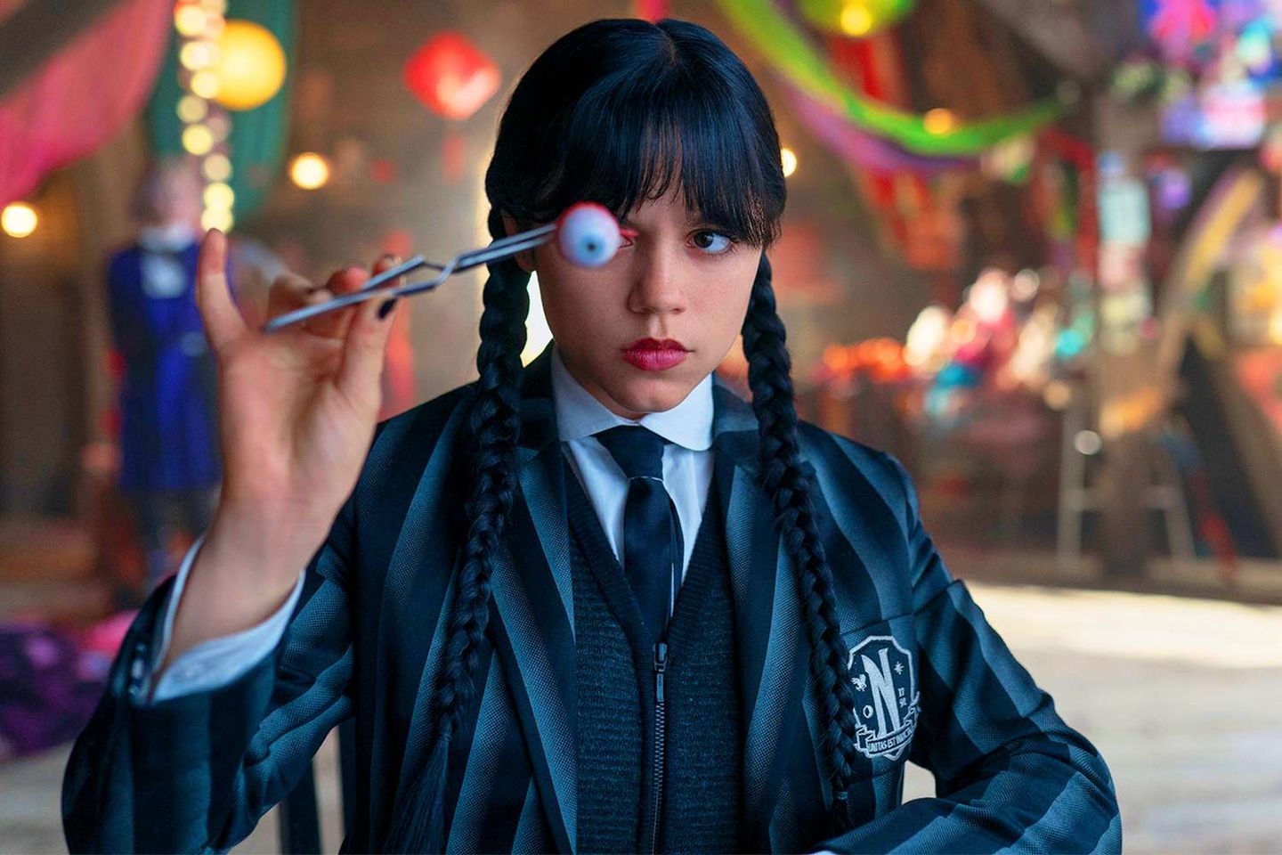 Young woman in dark school uniform with braids holds a fake eyeball with tongs, deadpan expression, colorful backdrop.