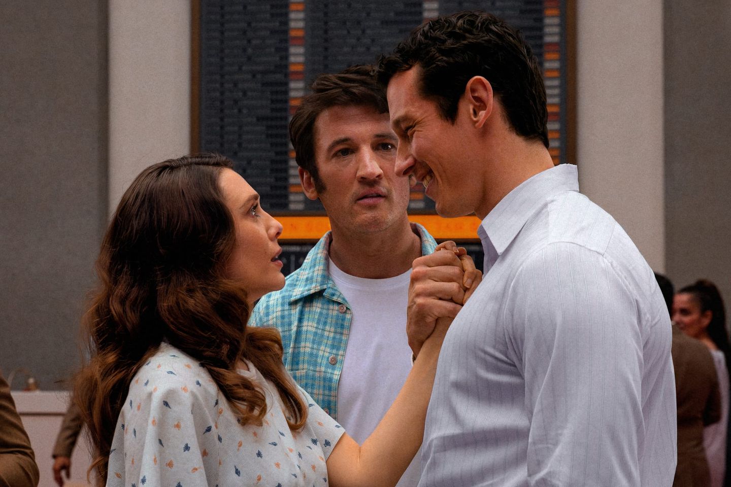 Three people in a train station: a woman and man dance closely while another man watches with a concerned expression.