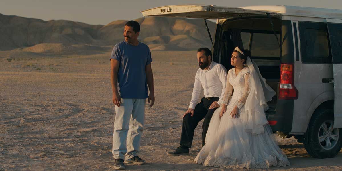 Three people in a desert: a man in blue shirt standing, a couple in wedding attire sitting in the back of a white van.