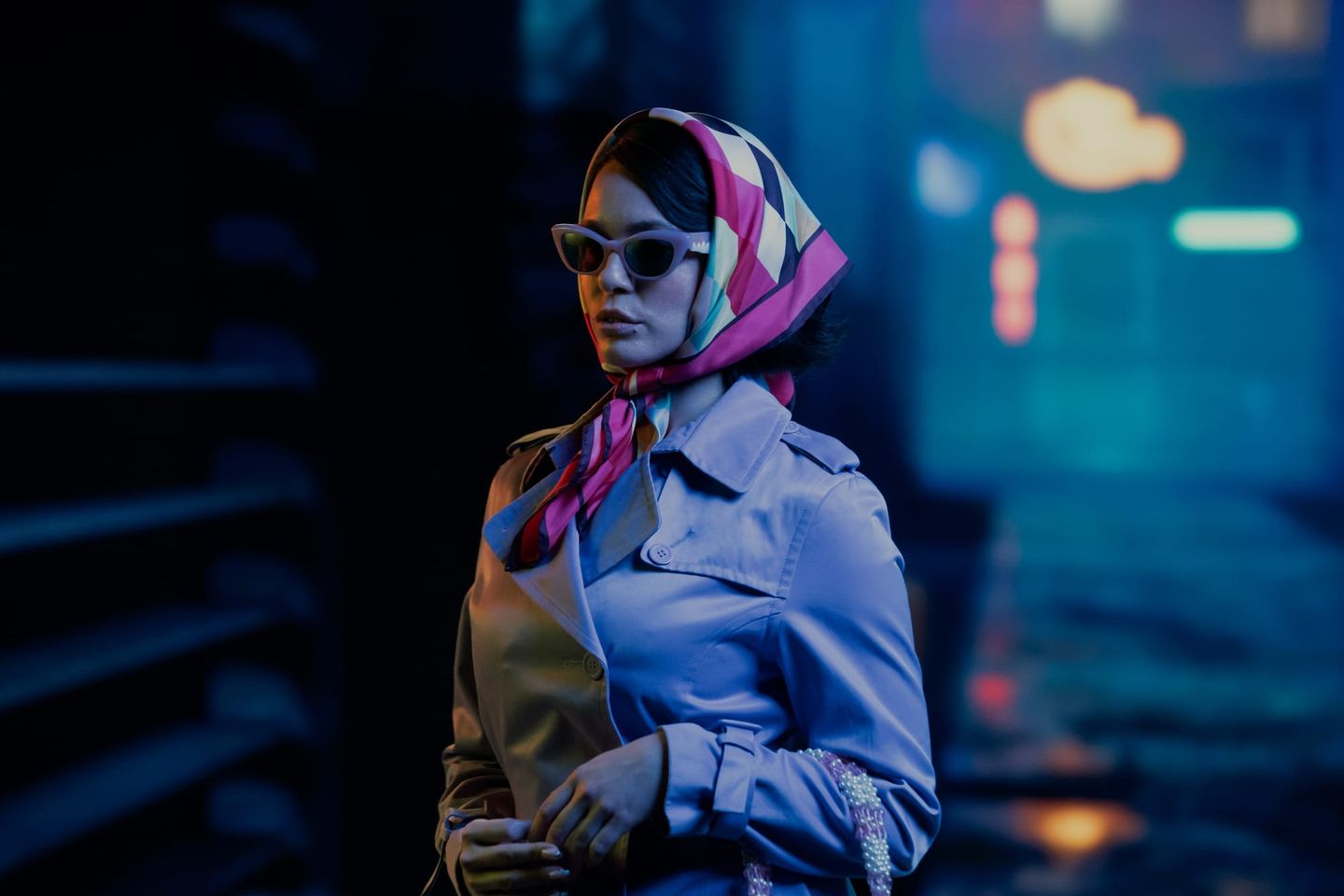 Woman in sunglasses, pink geometric headscarf, and trench coat poses in moody blue neon-lit urban night setting.