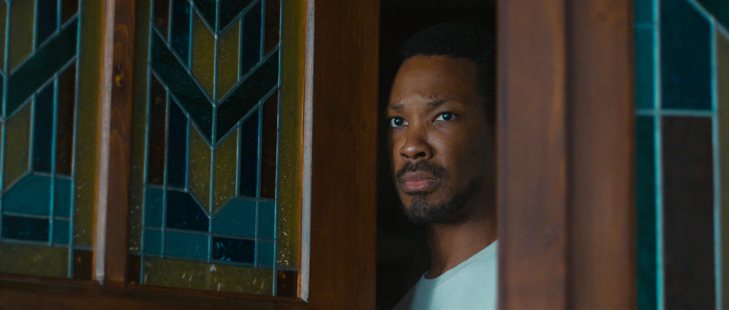 Man with short hair and beard peering cautiously through a doorway beside a geometric stained glass window in dim lighting.