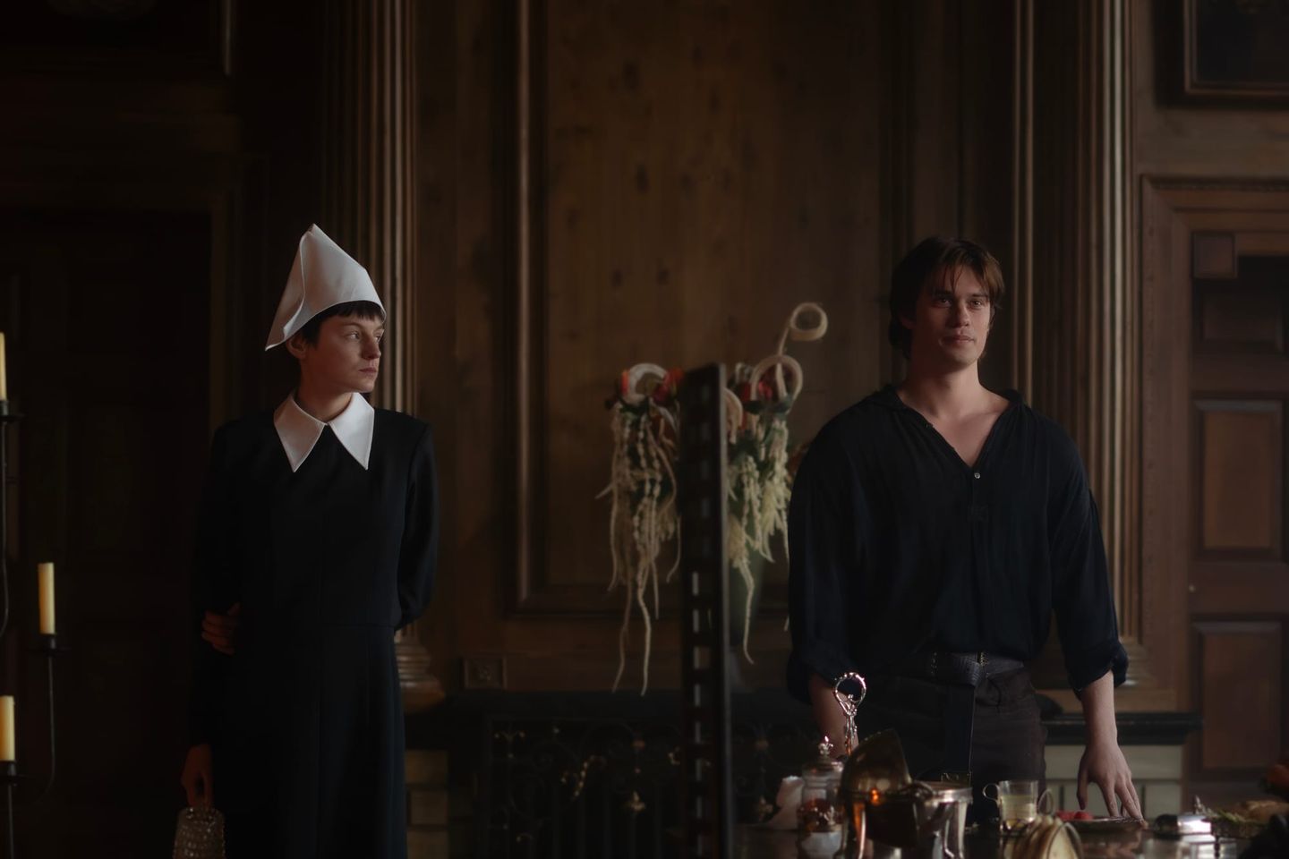 Two people in a dimly lit wood-paneled room; one wears a black dress with white collar and white pointed cap, the other a dark loose shirt.