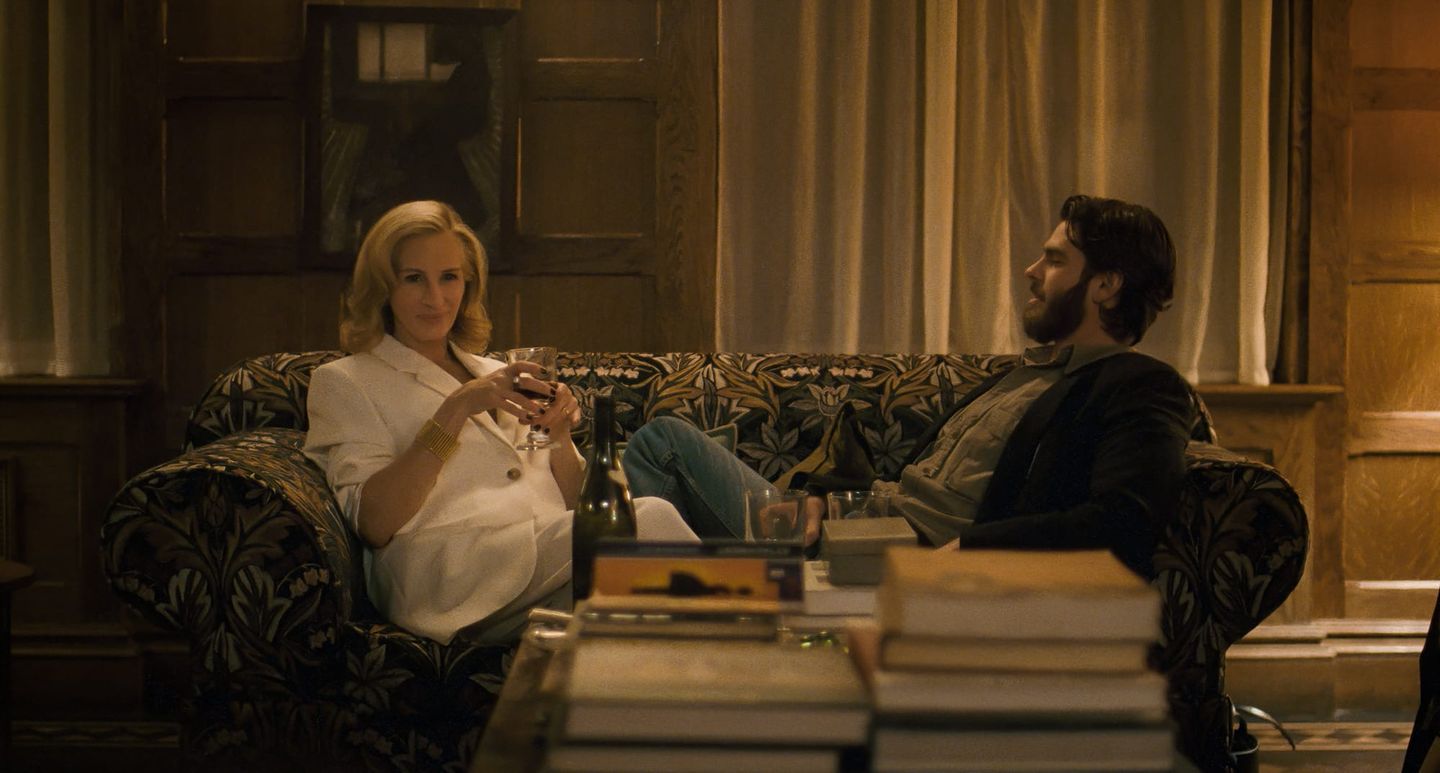 Two people sitting on a floral couch sharing drinks in a warmly lit wood-paneled room, with books and a wine bottle nearby.