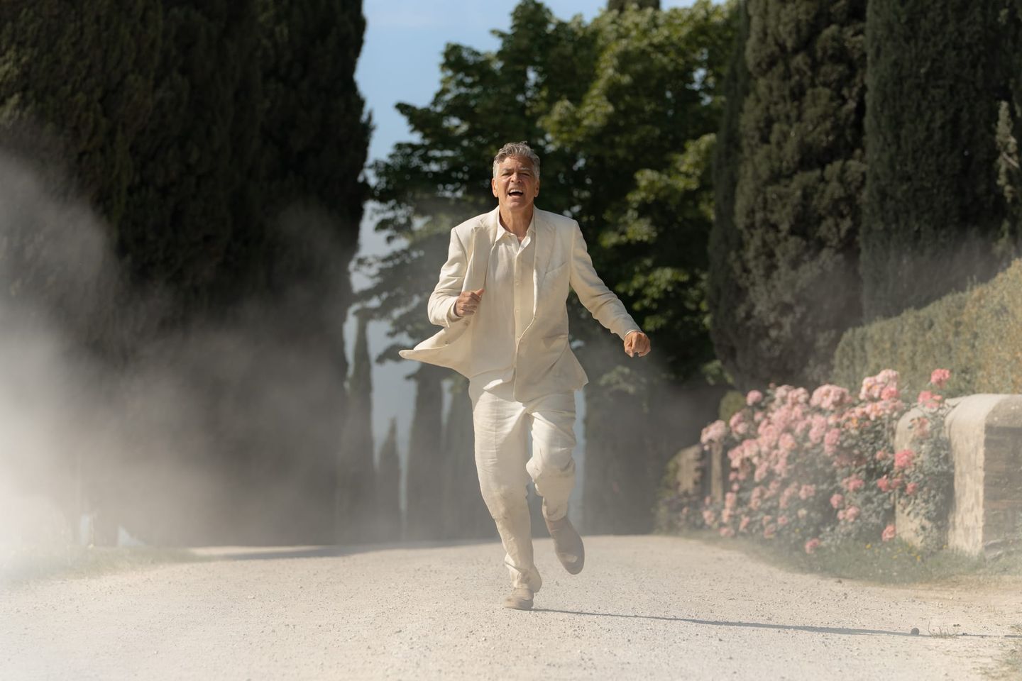 Man in white linen suit running urgently down a dusty cypress-lined path with pink roses and dust cloud behind him.