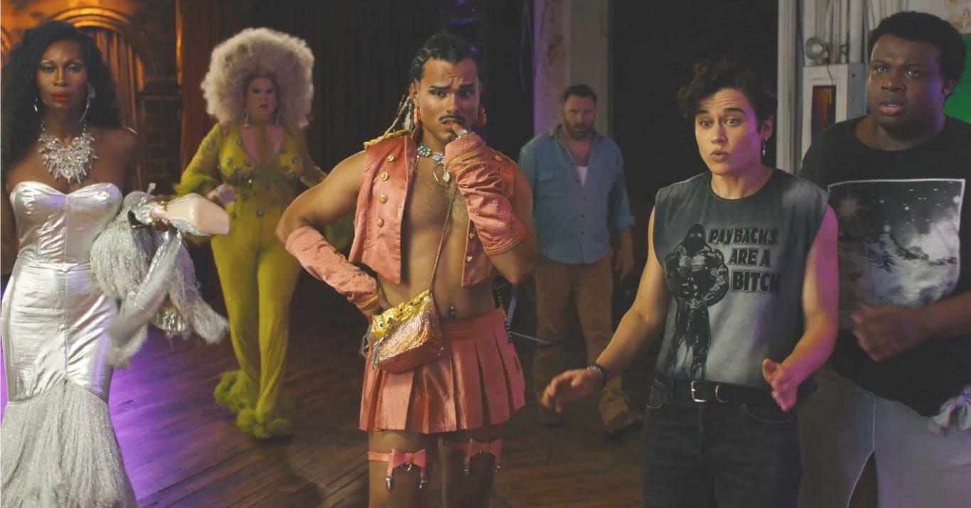 Six people in a colorful scene: drag queens in silver and yellow gowns, a person in pink skirt and open jacket, others in casual wear.