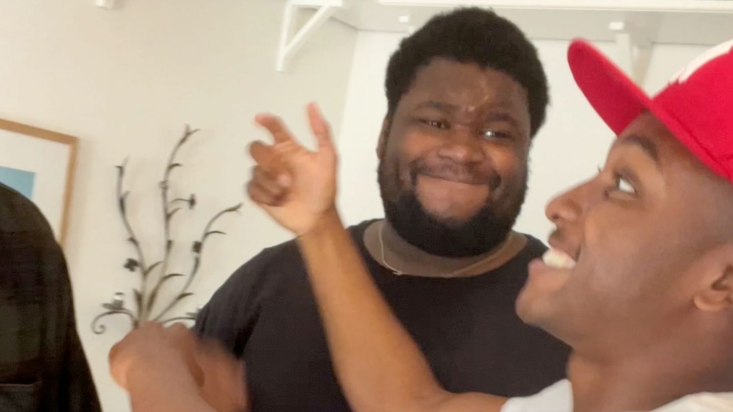 Two Black men smiling and laughing together indoors; one wears a black shirt, the other a red cap, with a plant behind them.