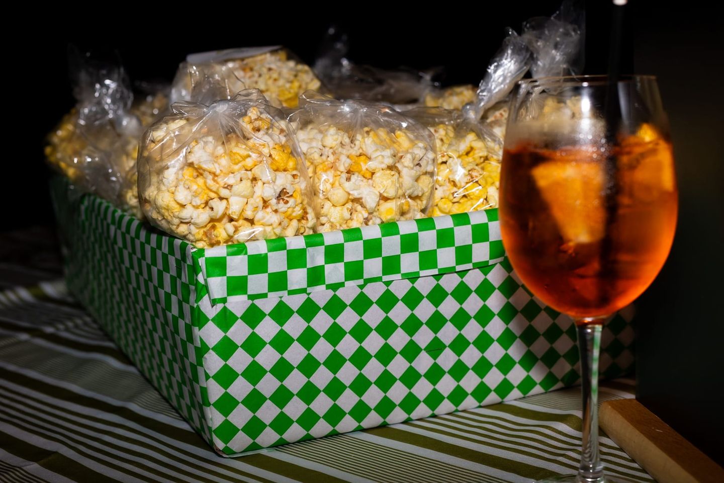 Bags of buttered popcorn in a green-and-white checkered box beside a glass of Aperol Spritz on a striped tablecloth.