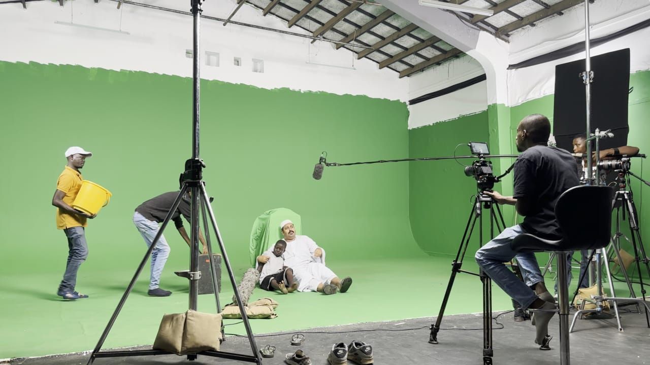 Film crew shooting a scene on a green screen set with two actors seated on the floor, a boom mic, cameras, and crew members.