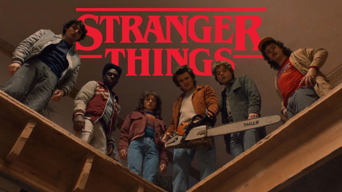 Stranger Things promotional still: six young cast members look down into a hole, one holding a Stihl chainsaw, with the show's red logo above.