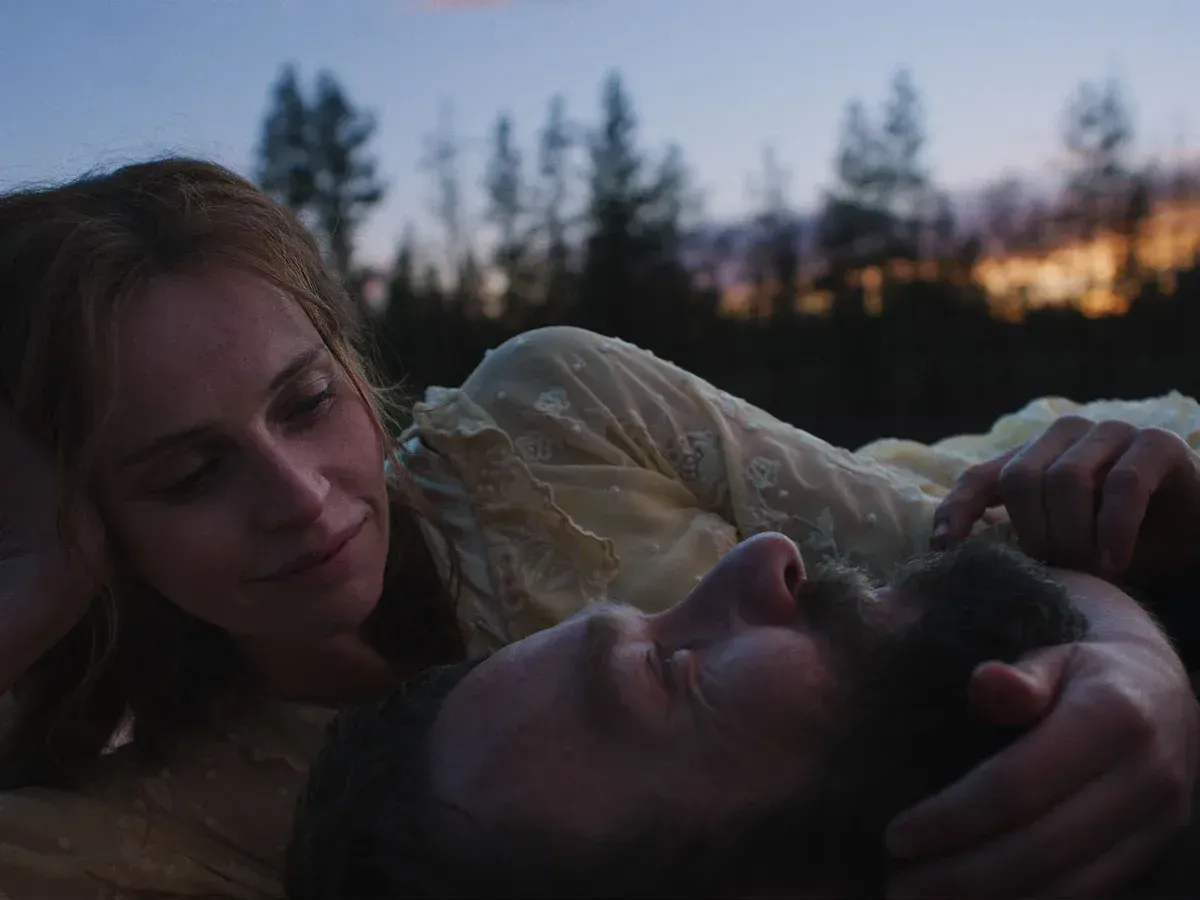 A woman gazes down tenderly at a man lying on his back outdoors at dusk, with pine trees and sunset glow in the background.
