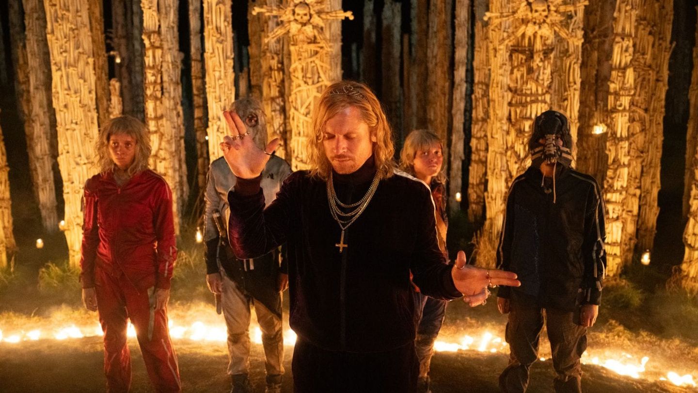 Five people stand in a dark forest with fire-lit ground and tall bone-decorated totems; center man wears black with cross necklace.