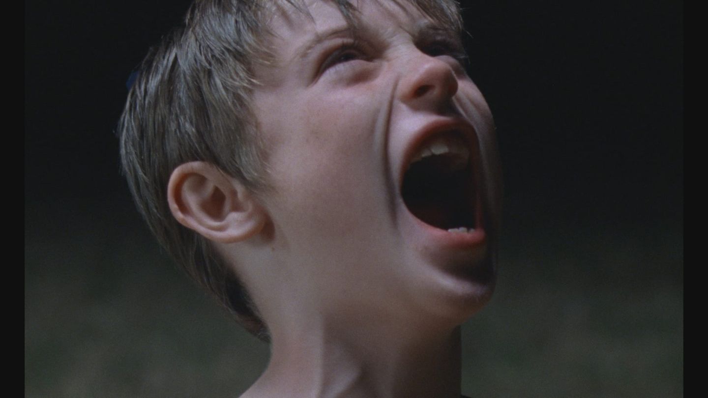 Close-up of a young boy with short blond hair screaming with his mouth wide open, head tilted back, against a dark background.