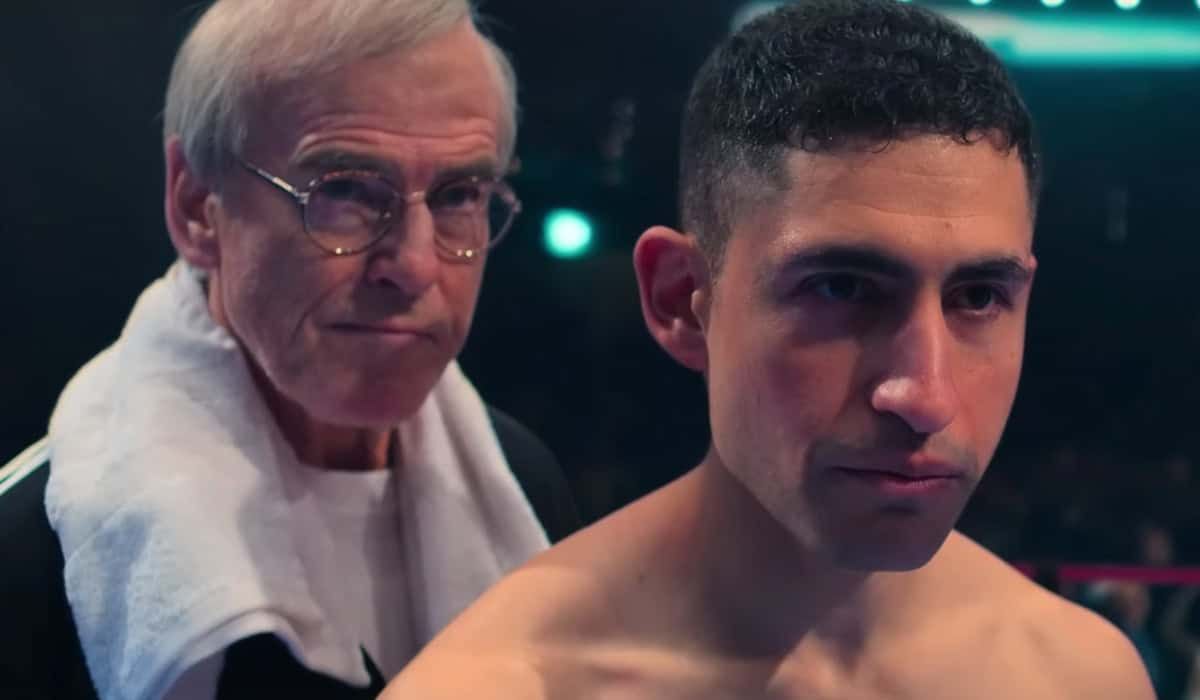 Older man with glasses and towel on shoulder stands behind shirtless young boxer in a dimly lit boxing ring setting.