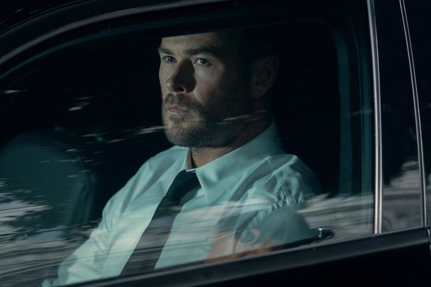 Man with beard in white dress shirt and black tie sitting in car, viewed through window with reflections, moody lighting.