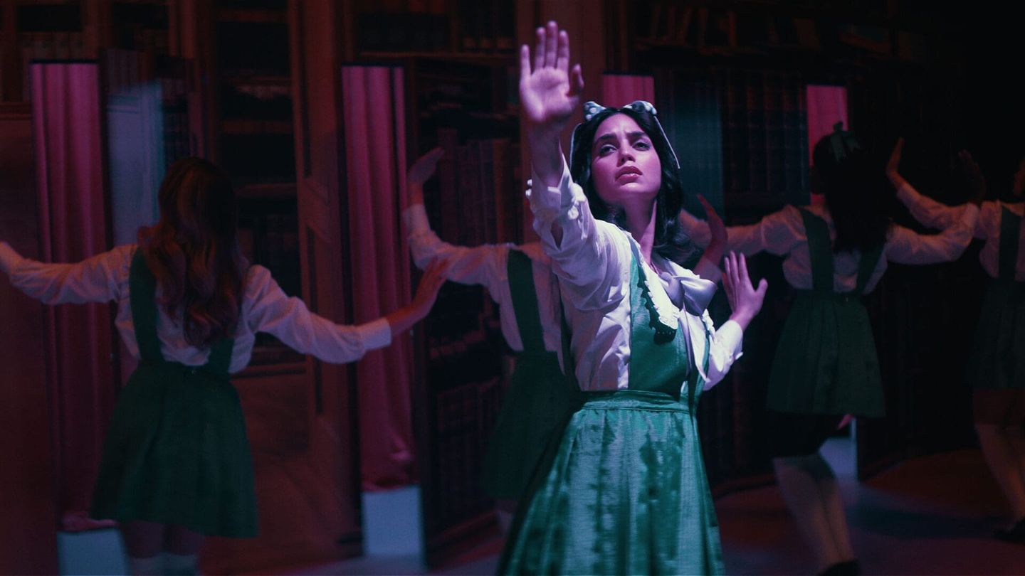 Young woman in green pinafore dress and white blouse dances with raised hands amid other dancers in a dimly lit library.
